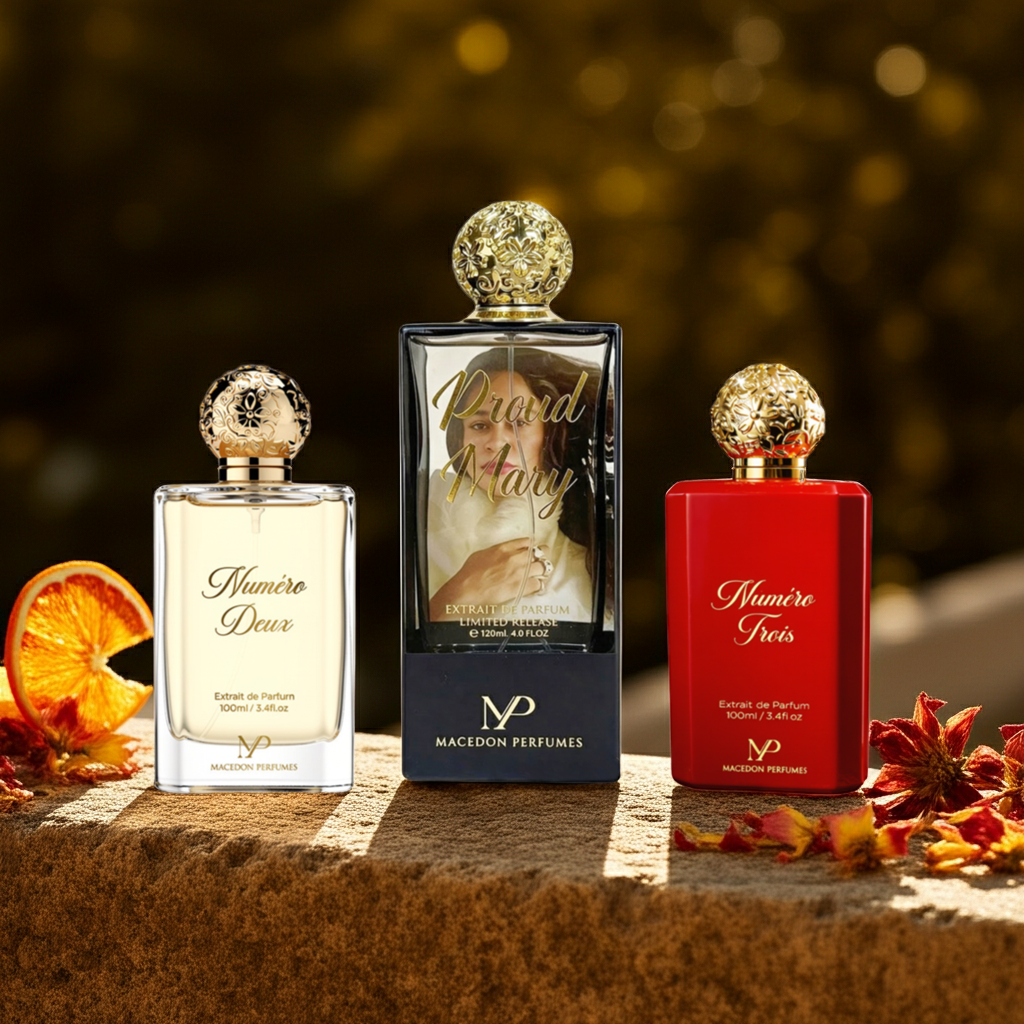 Three perfume bottles from Macaron Perfumes are displayed on a stone surface with autumn leaves and a dried orange slice. The center bottle is the featured 'Pour Mary' Extrait de Parfum, a rectangular glass bottle with a black base, gold cap, and a portrait of Mary on the front.